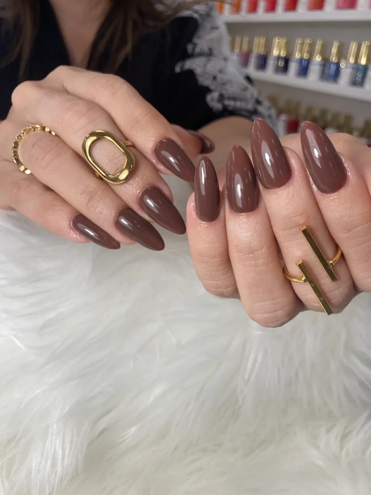 Long chocolate brown gel almond nails, rings on, polish wall behind